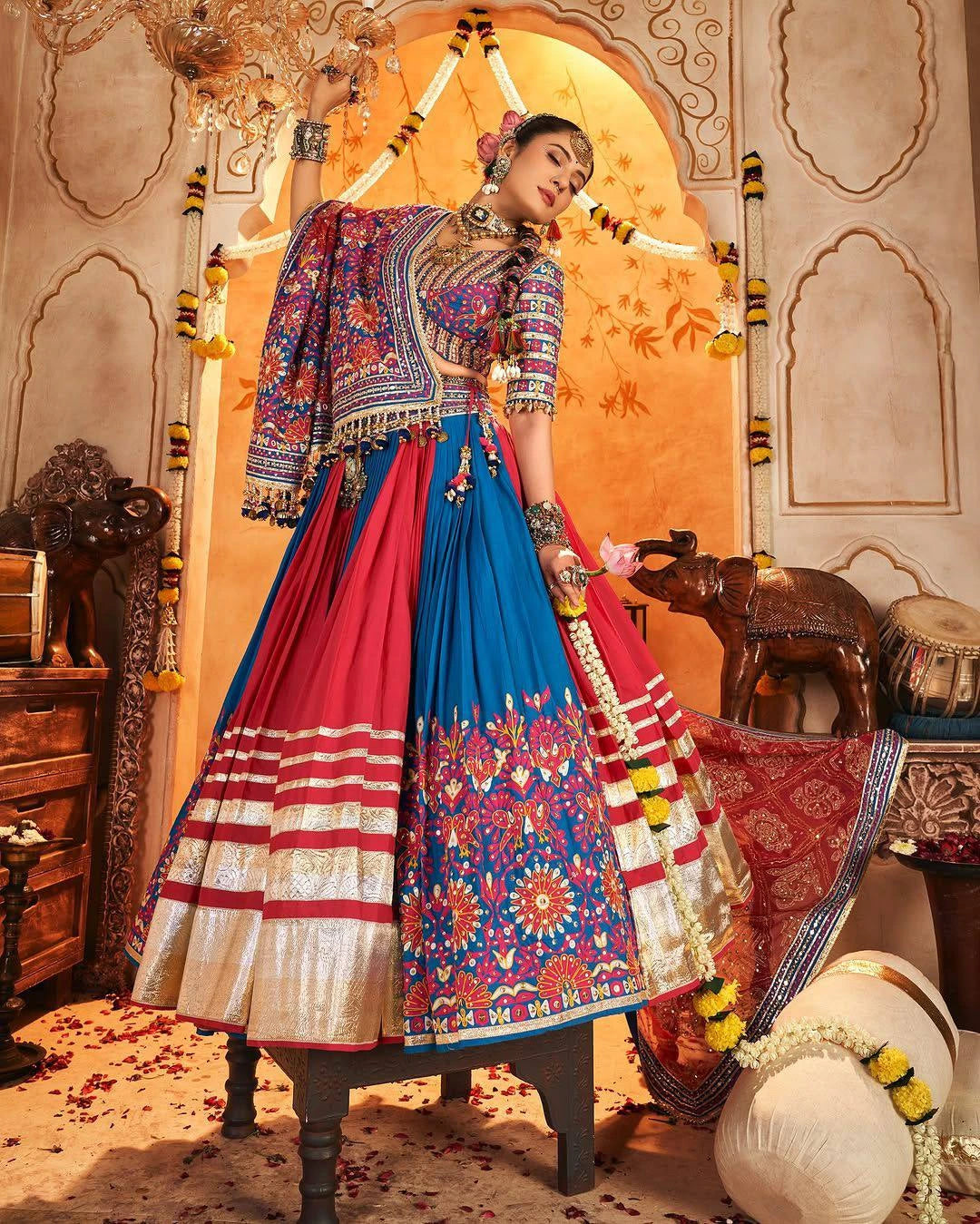 Woman in traditional Indian attire with a colorful blouse and red and blue lehenga, standing in an ornate decorated room.