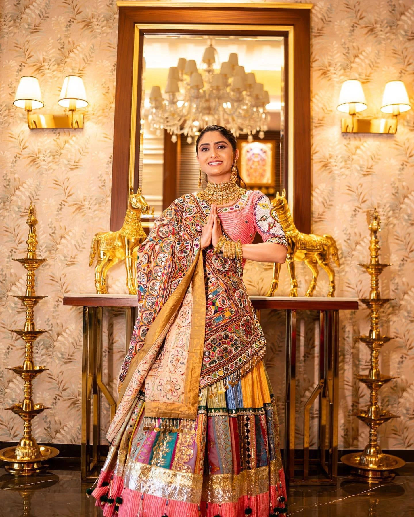 Woman in traditional multicolored outfit standing in a decorated room with chandelier and wall lights.