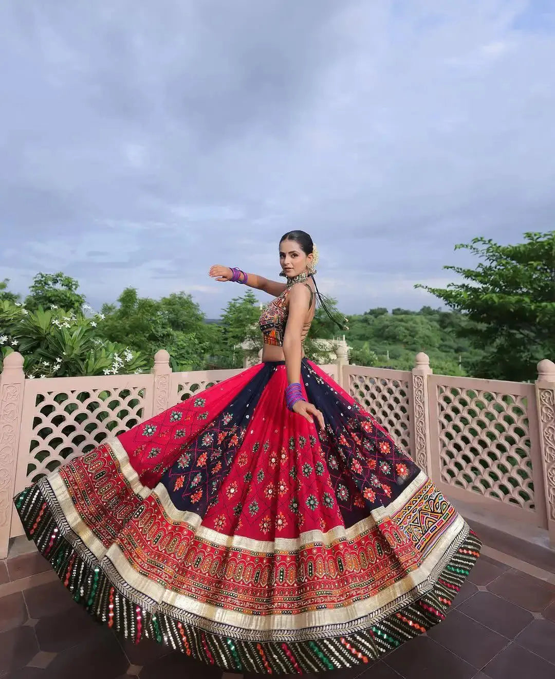 Woman in a colorful traditional outfit with a patterned skirt, standing outdoors with trees in the background.