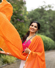 Woman in an orange saree with a pink blouse standing outdoors with greenery in the background