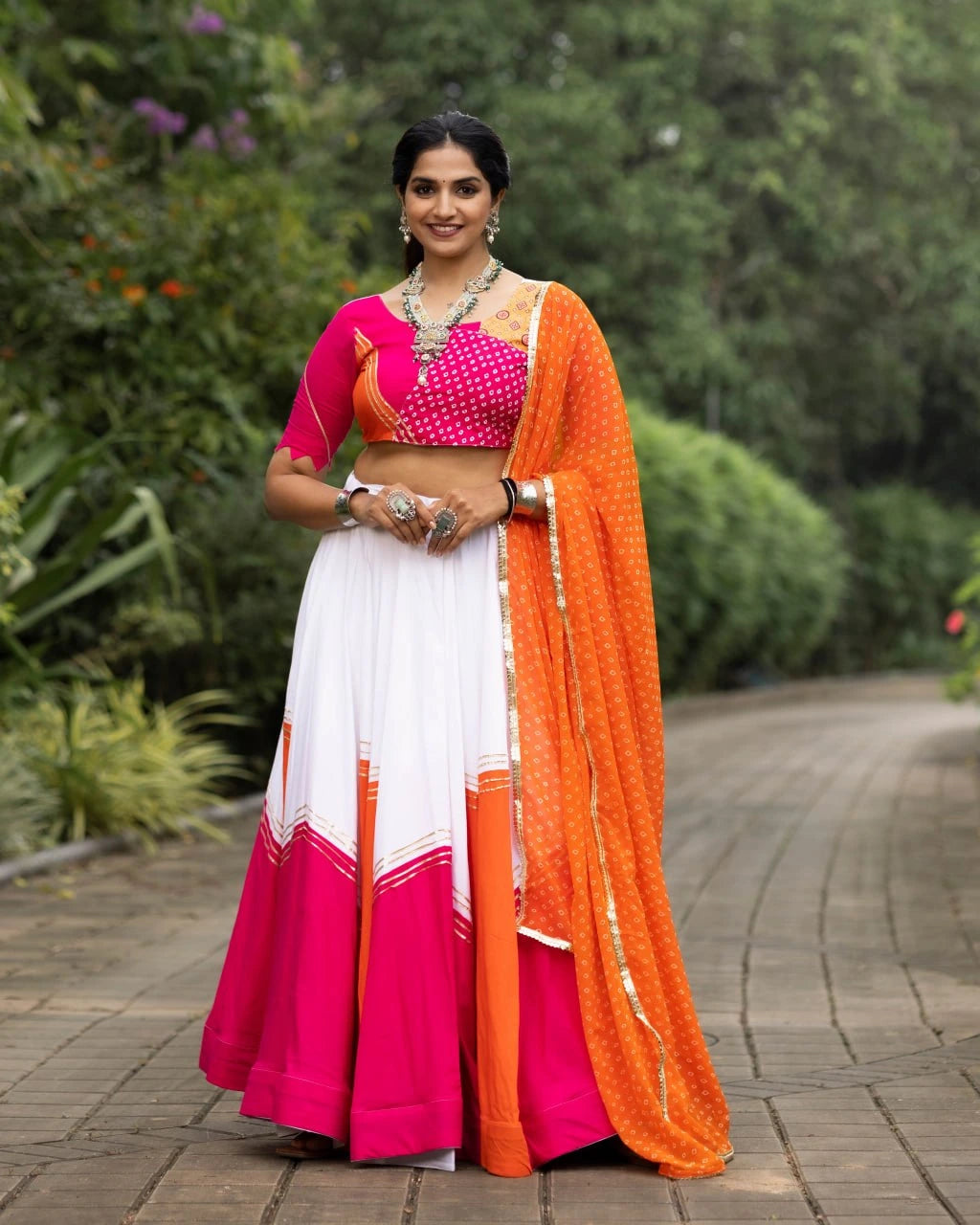 Woman in a pink and orange traditional outfit standing outdoors with greenery in the background