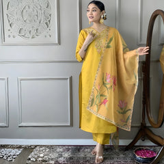 Woman in a yellow traditional outfit with floral patterns standing against a decorative wall.