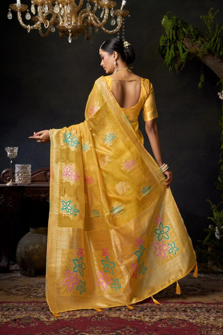 Woman in a yellow saree with floral patterns standing in a room with a chandelier.