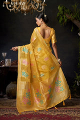 Woman in a yellow saree with floral patterns standing in a room with a chandelier.