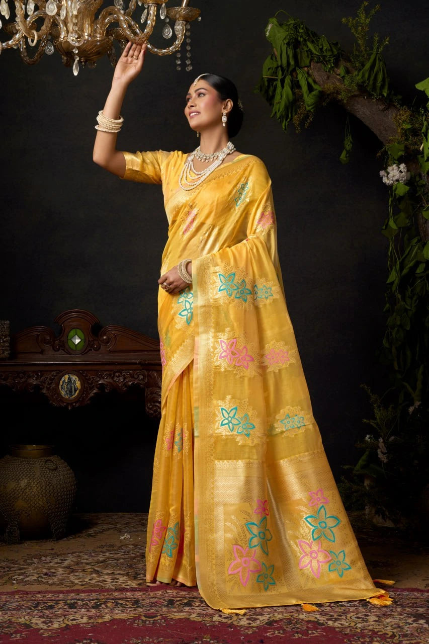 Woman in a yellow saree with floral patterns standing indoors against a dark background.