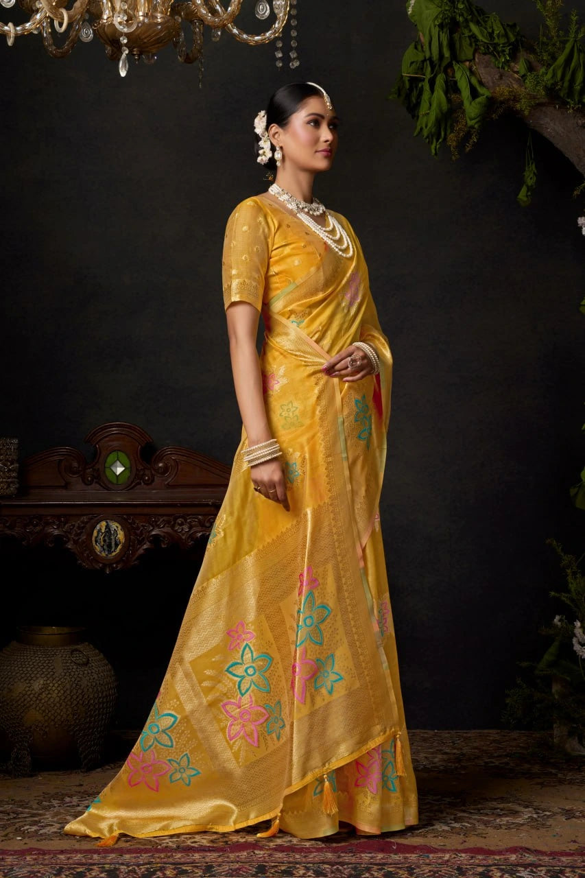 Woman in a yellow saree with floral patterns standing against a dark background
