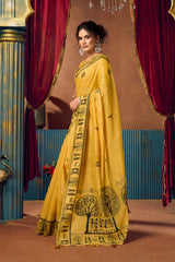 Woman in a yellow saree with black patterns standing in front of red curtains.