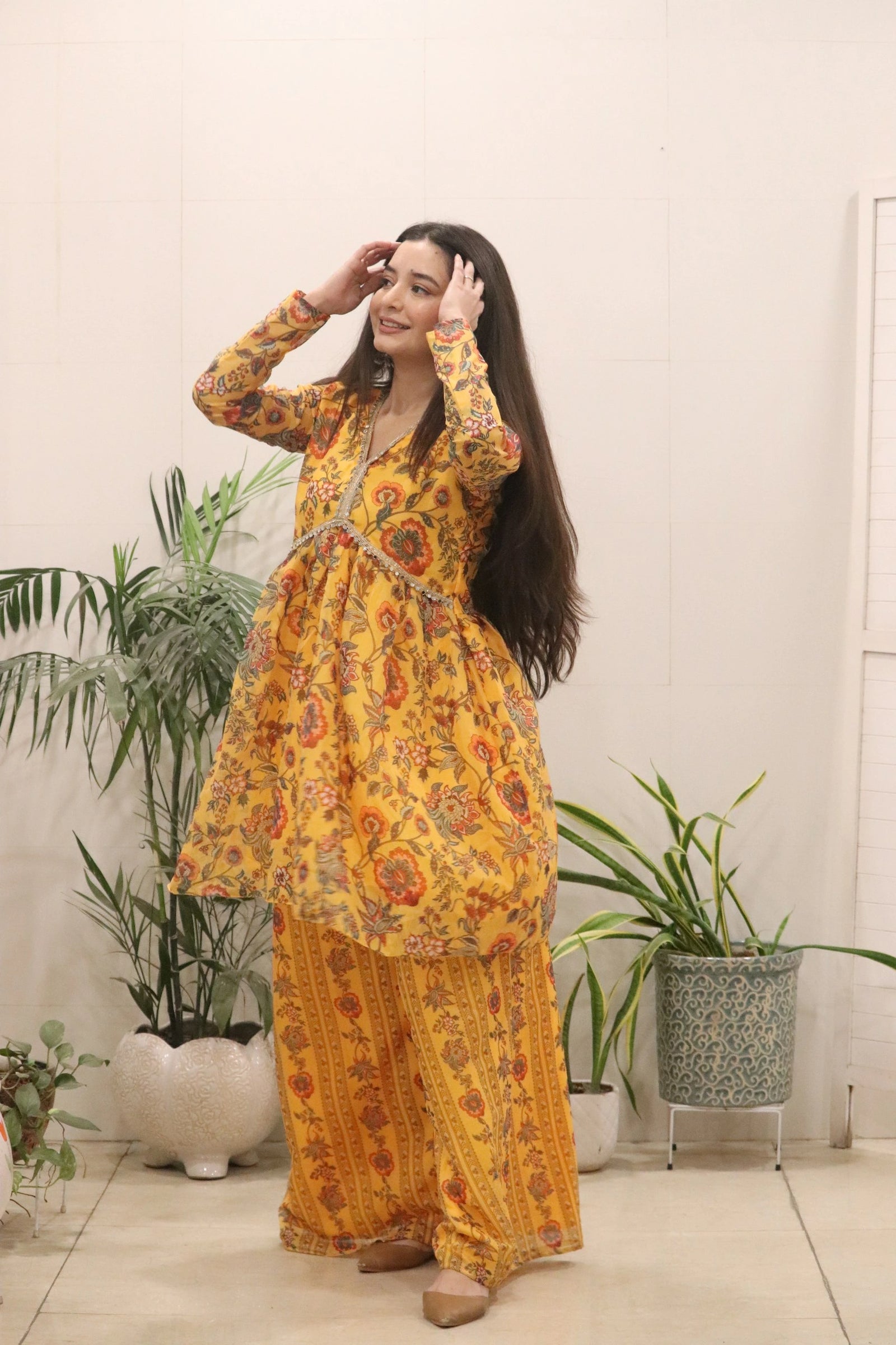 Woman wearing a yellow floral outfit standing indoors with plants in the background