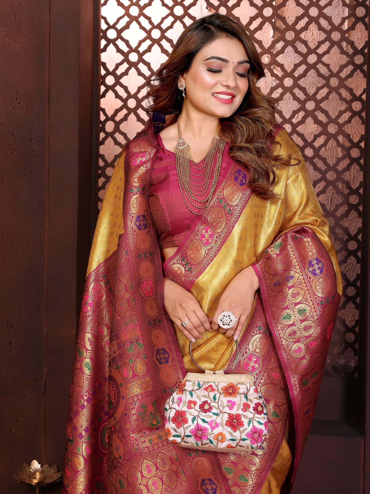 Woman in traditional maroon and gold saree with jewelry and floral clutch against a decorative wall.
