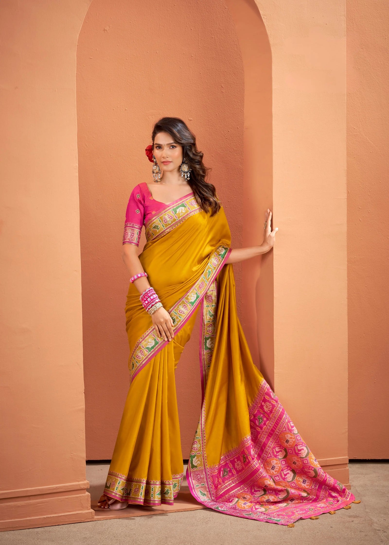 Woman wearing a yellow and pink saree with a decorative border against a peach-colored wall.