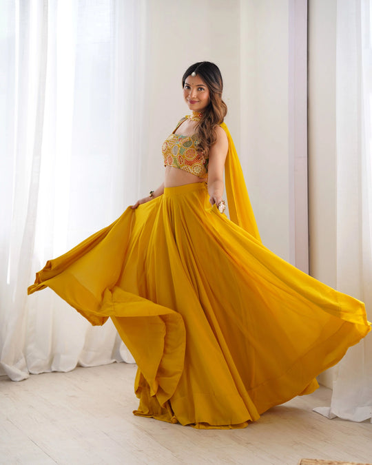 Woman in a yellow traditional outfit with a floral top, standing against white curtains.