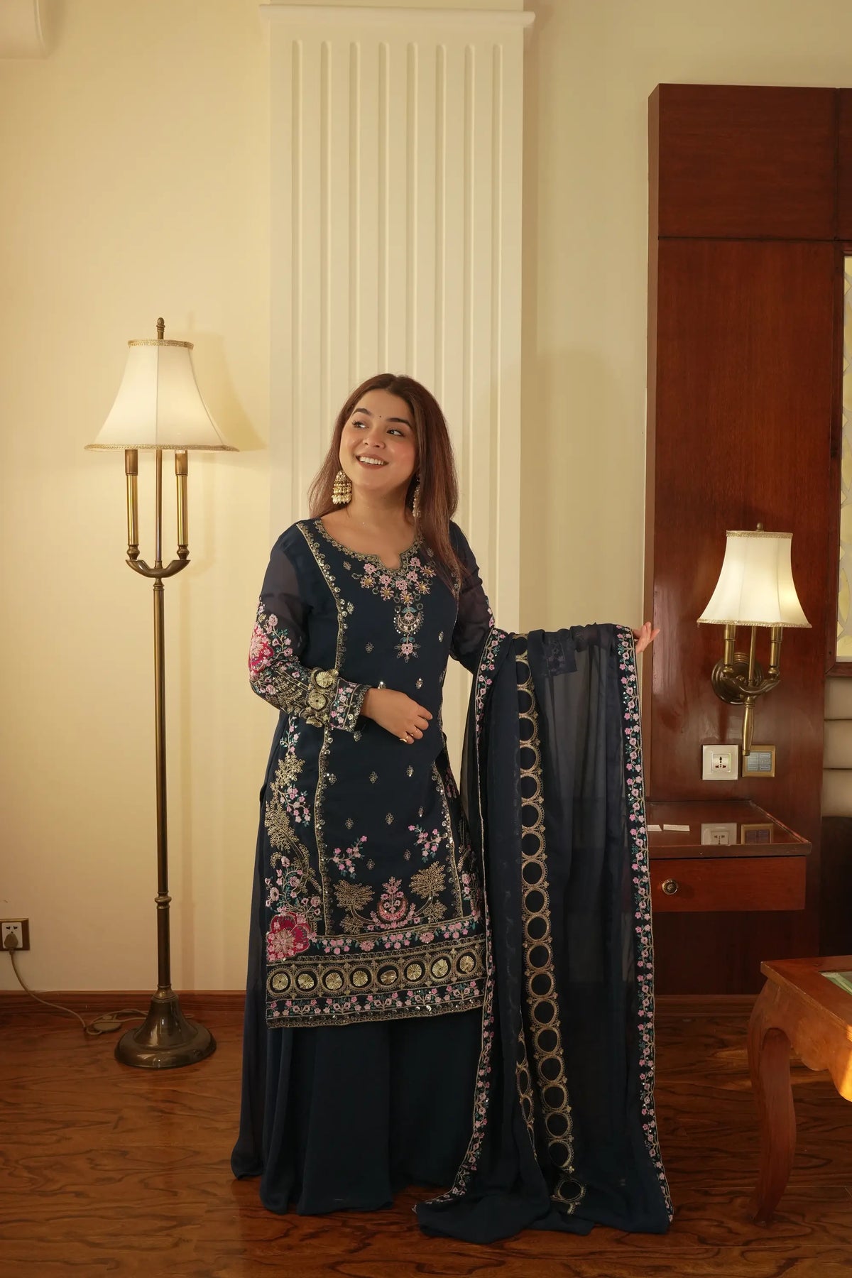 Woman in a dark blue embroidered outfit holding a matching dupatta in a room with a lamp and wooden furniture.