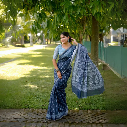 A woman models a striking Navy Blue Soft Cotton Saree featuring an all-over digital block print of traditional white motifs. She wears a light blue patterned blouse and silver jhumka earrings, posing on a path in a green outdoor setting.