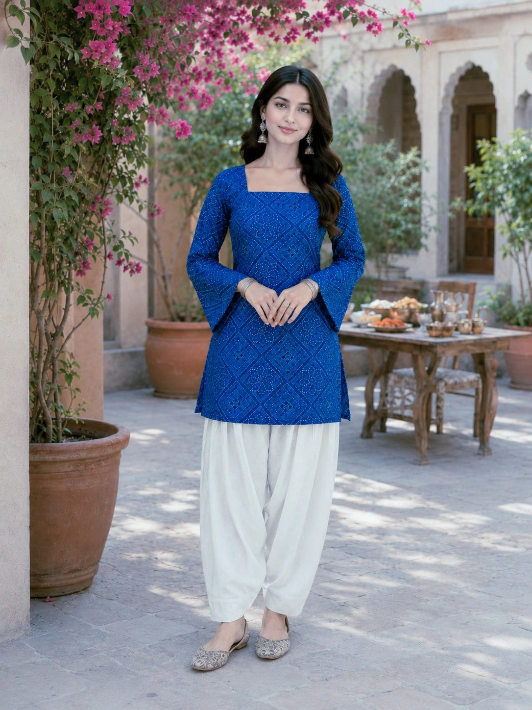 Woman in a blue top and white pants standing outdoors with flowers and a table in the background