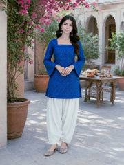 Woman in a blue top and white pants standing outdoors with flowers and a table in the background