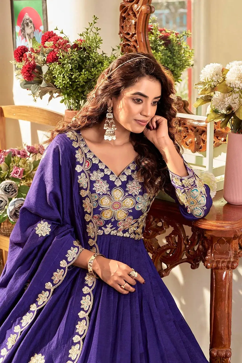 Woman in a blue embroidered traditional outfit sitting in a decorated room with flowers and furniture.