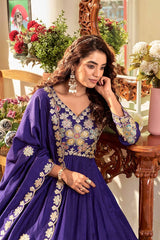 Woman in a blue embroidered traditional outfit sitting in a decorated room with flowers and furniture.