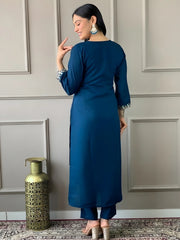 Woman in a blue outfit standing against a gray paneled wall with a decorative vase.