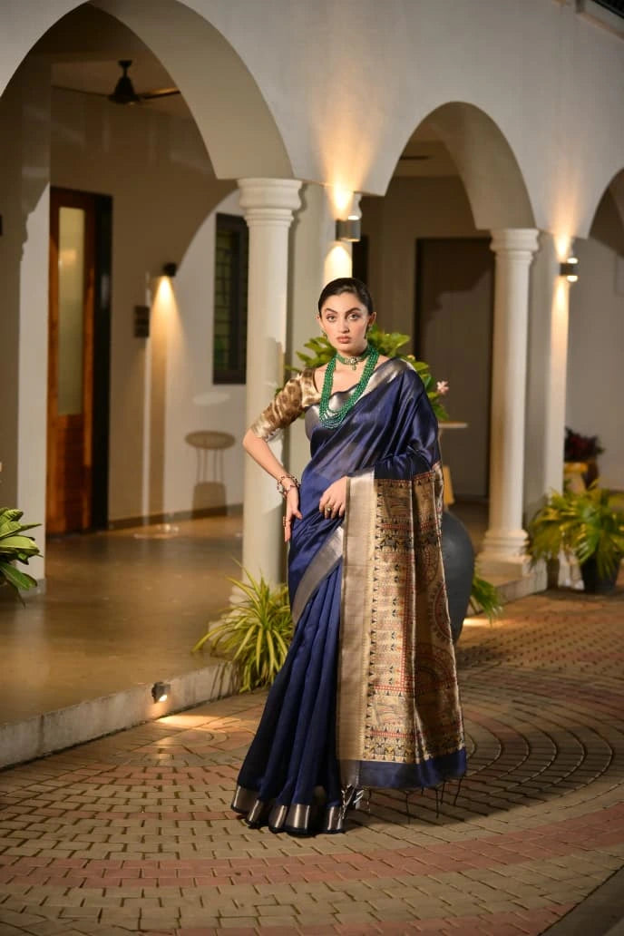 Woman in a blue saree with a patterned border standing in an indoor setting with columns and plants.