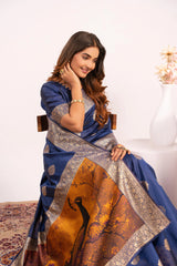 Woman wearing a blue saree with a peacock design, sitting on a white chair.