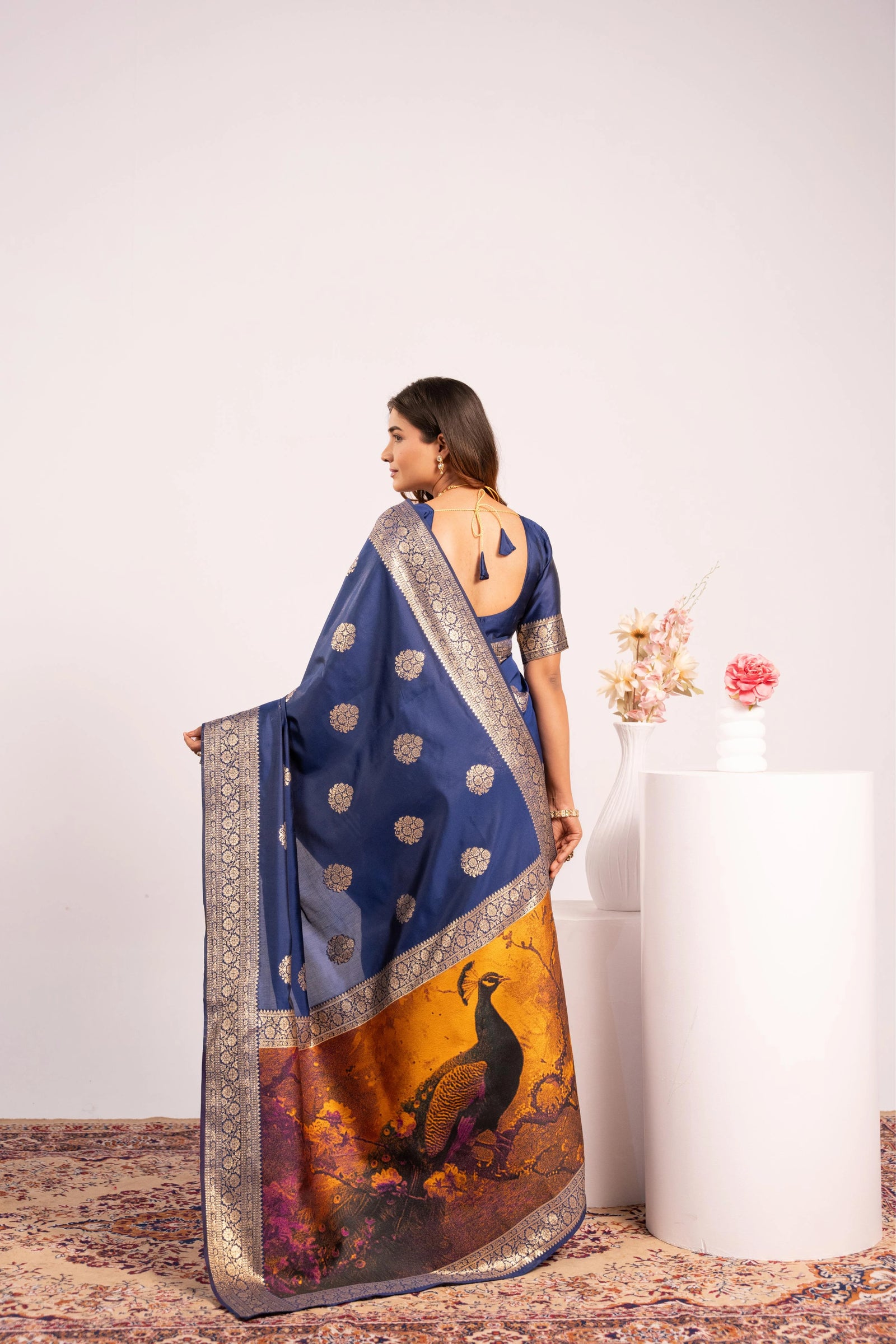 Woman wearing a blue saree with a peacock design, standing against a white wall.