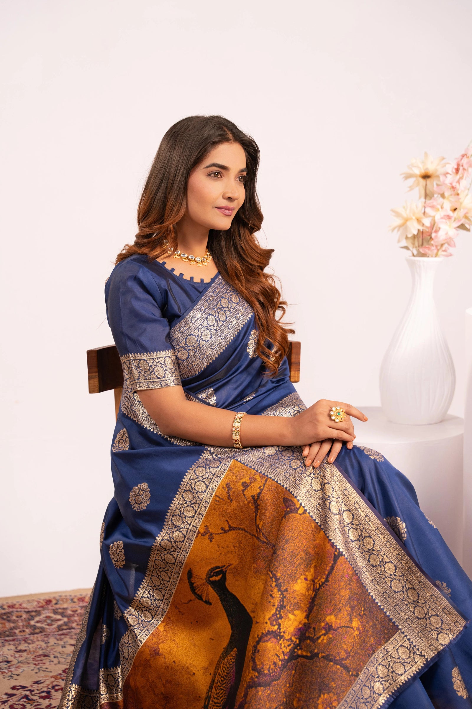 Woman wearing a blue saree with a peacock design, sitting on a chair against a white background.