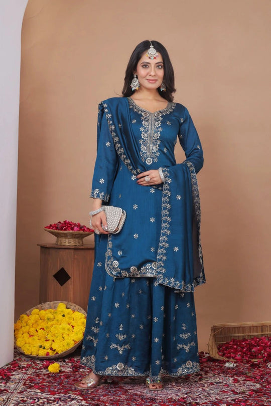 Woman in a blue embroidered traditional outfit standing in a decorated room with flowers and a basket.