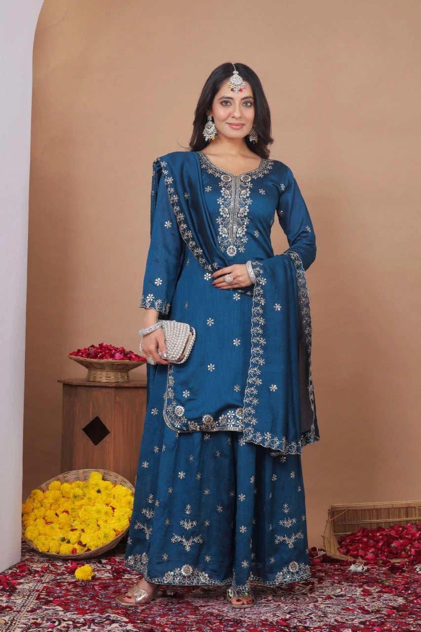 Woman in a blue embroidered traditional outfit standing in a decorated room with flowers and a basket.