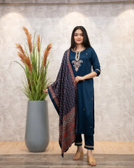 Woman in a blue traditional outfit standing next to a plant against a neutral wall.