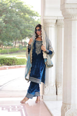 Woman in traditional blue outfit with embroidery standing in a decorative setting.