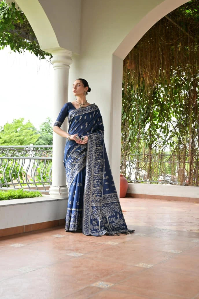 Woman in a blue saree standing on a wooden floor with greenery in the background