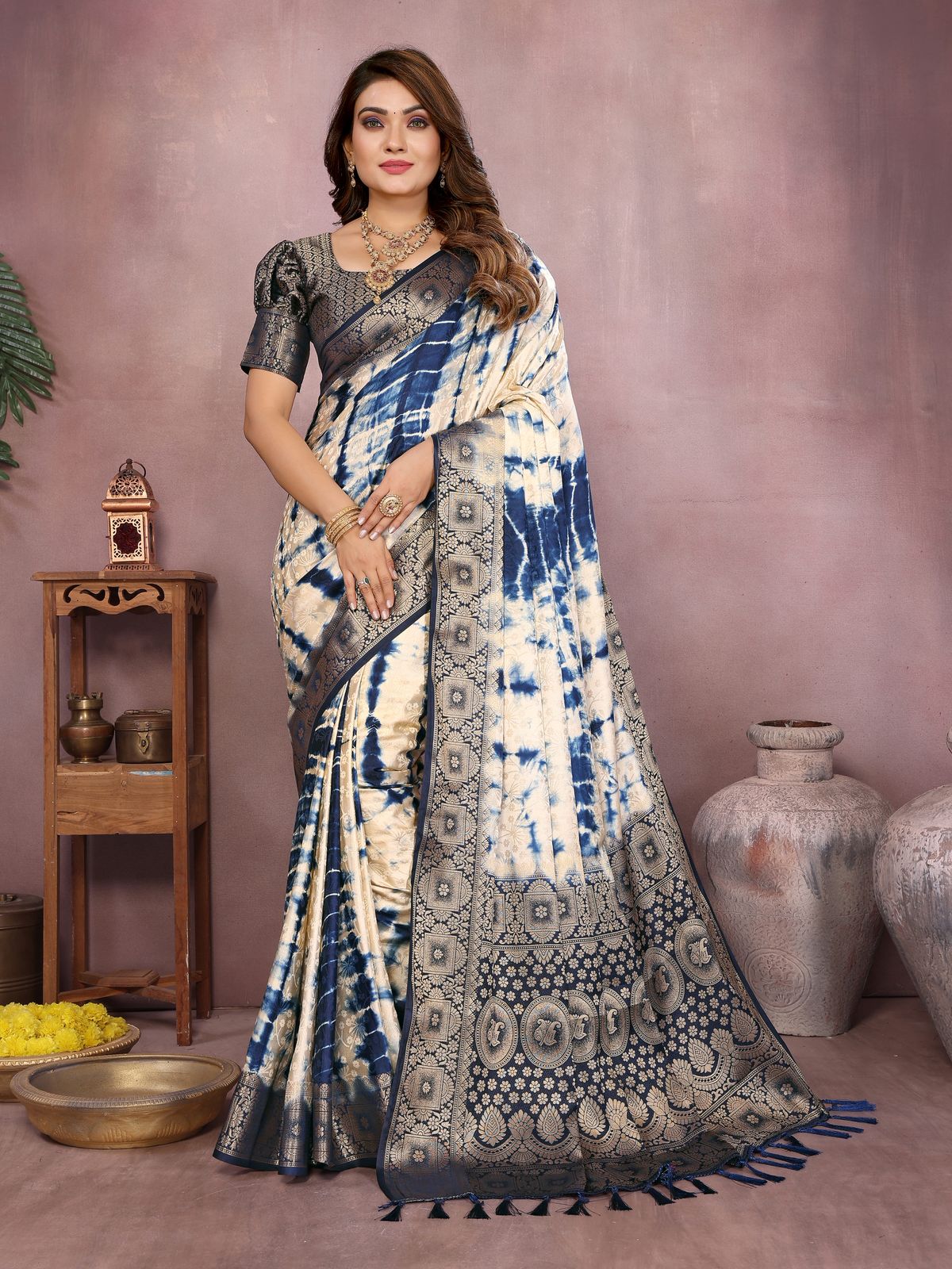 Woman wearing a blue and white saree with a patterned border against a pinkish-brown background.