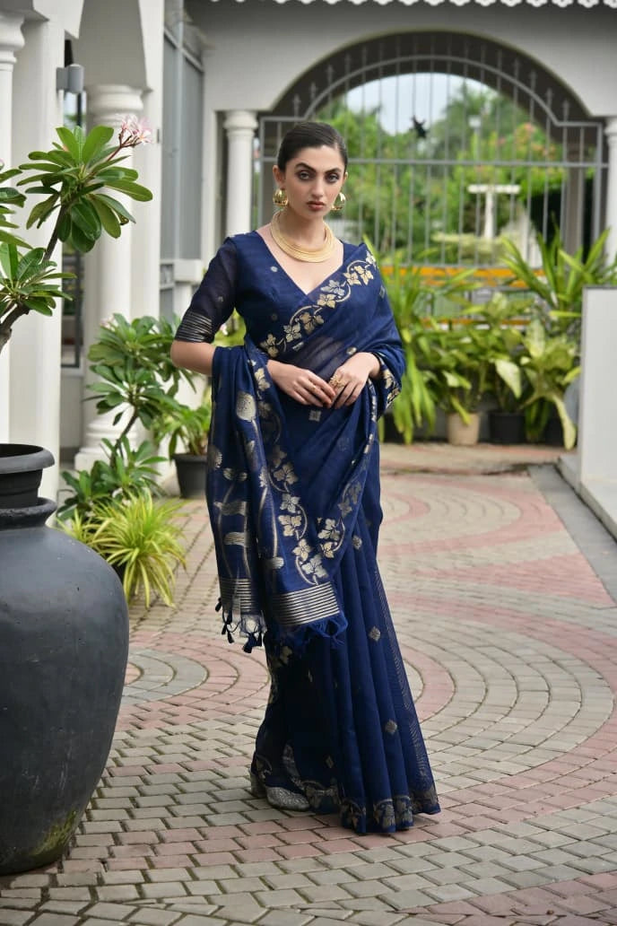 Woman in a blue saree standing outdoors with plants and a building in the background