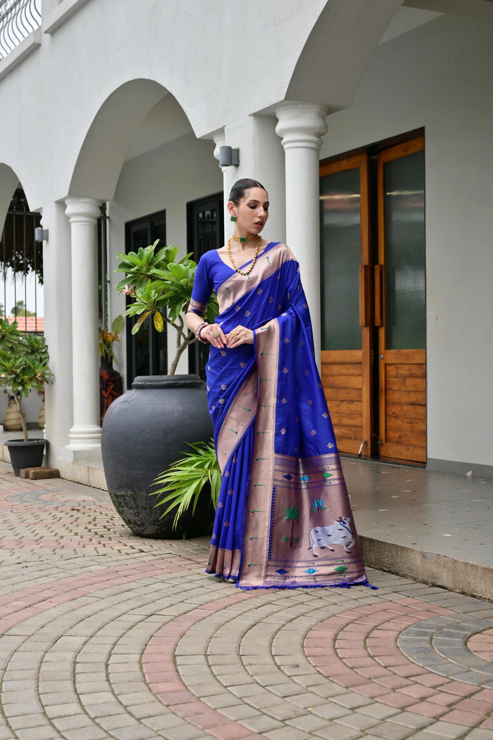 Woman in a blue saree standing in front of a building with columns and plants.
