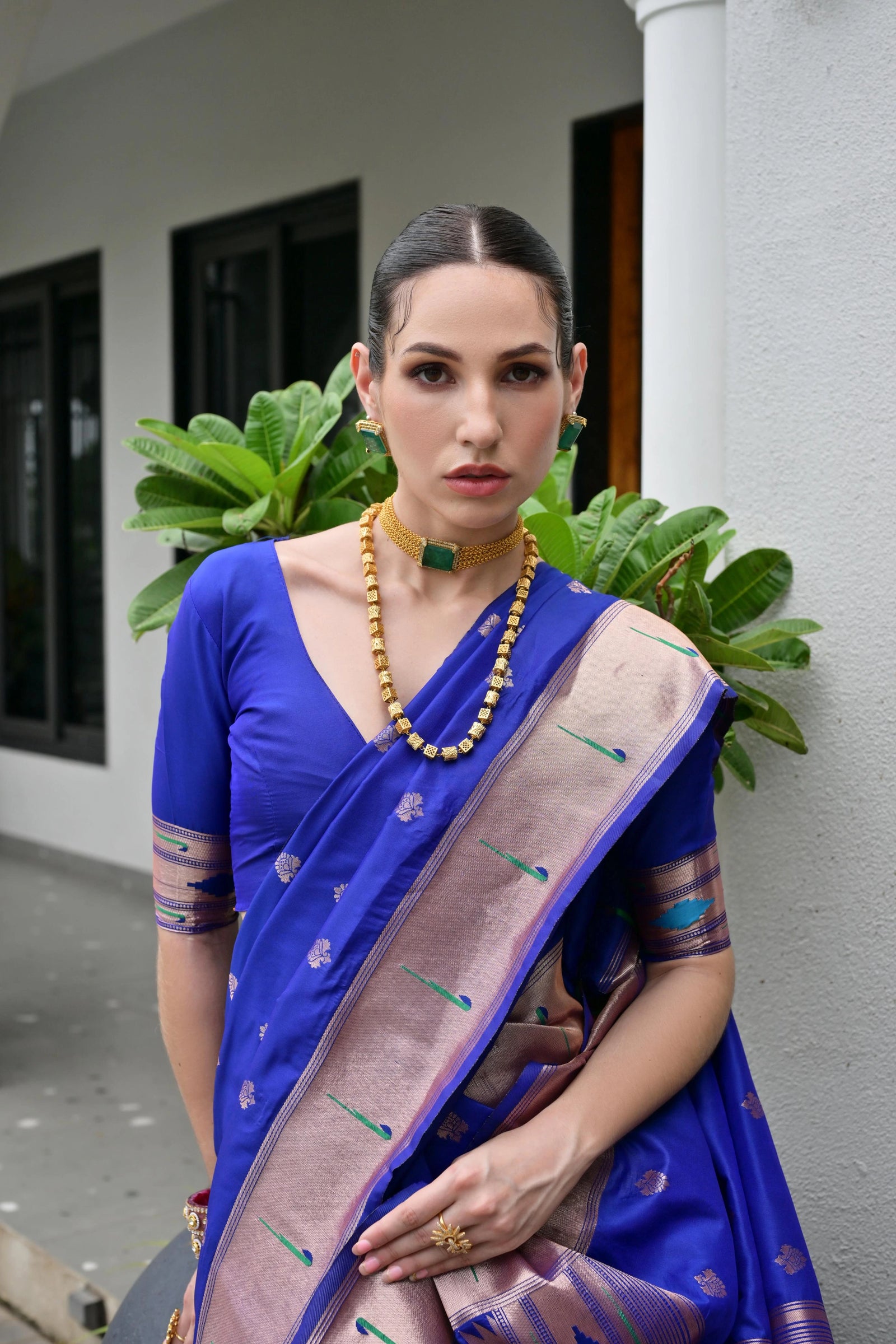 Woman in a blue saree with a purple border standing against a white wall with green plants.