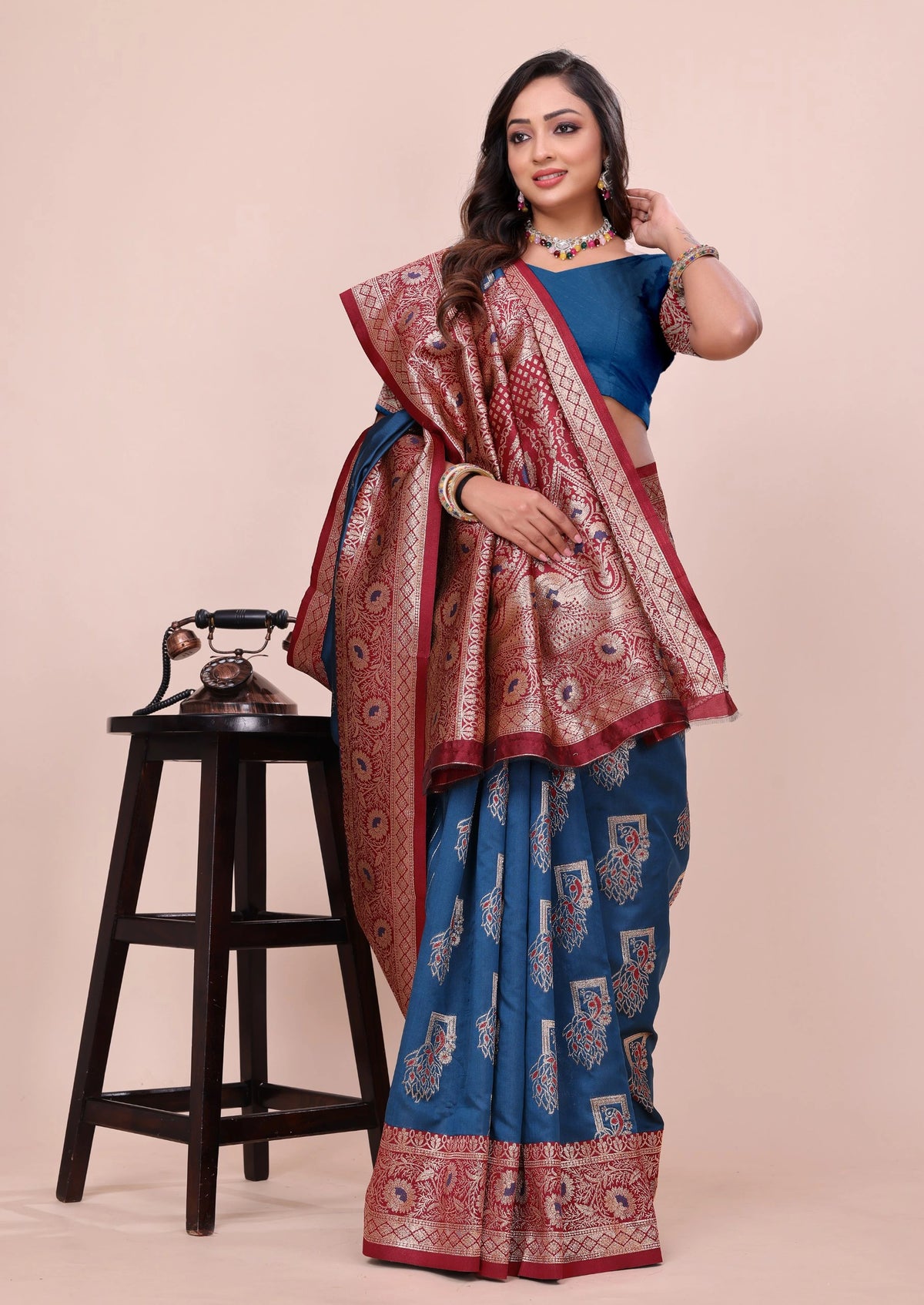 Woman wearing a blue saree with a red border, standing next to a wooden stool on a beige background.