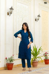Woman in a blue outfit standing in a decorated room with plants and wall lights.