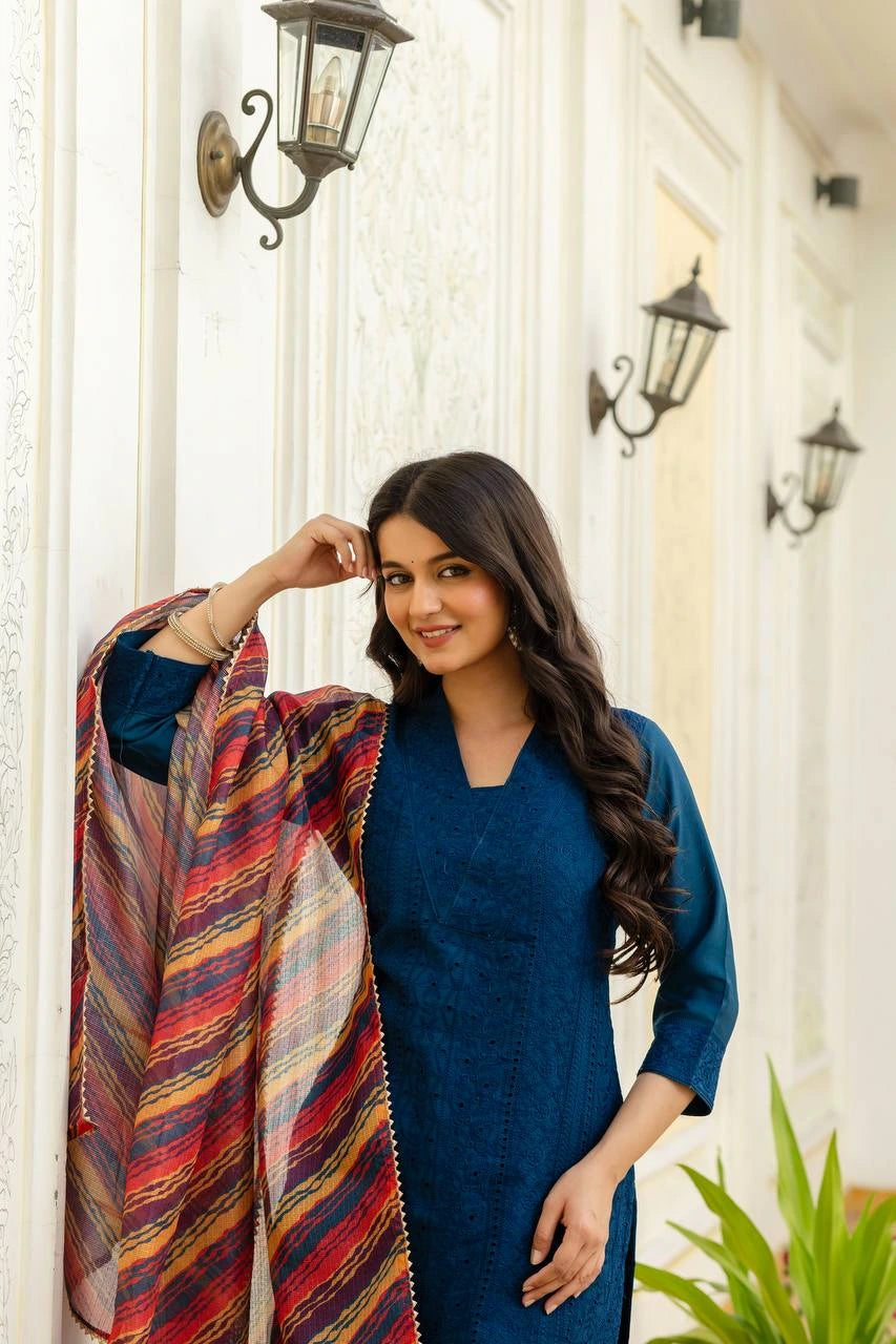 Woman in a blue outfit holding a colorful shawl in front of a white wall with decorative lights.