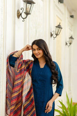 Woman in a blue outfit holding a colorful shawl in front of a white wall with decorative lights.