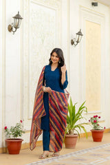 Woman in a blue outfit with a colorful scarf standing in a decorated room with plants.