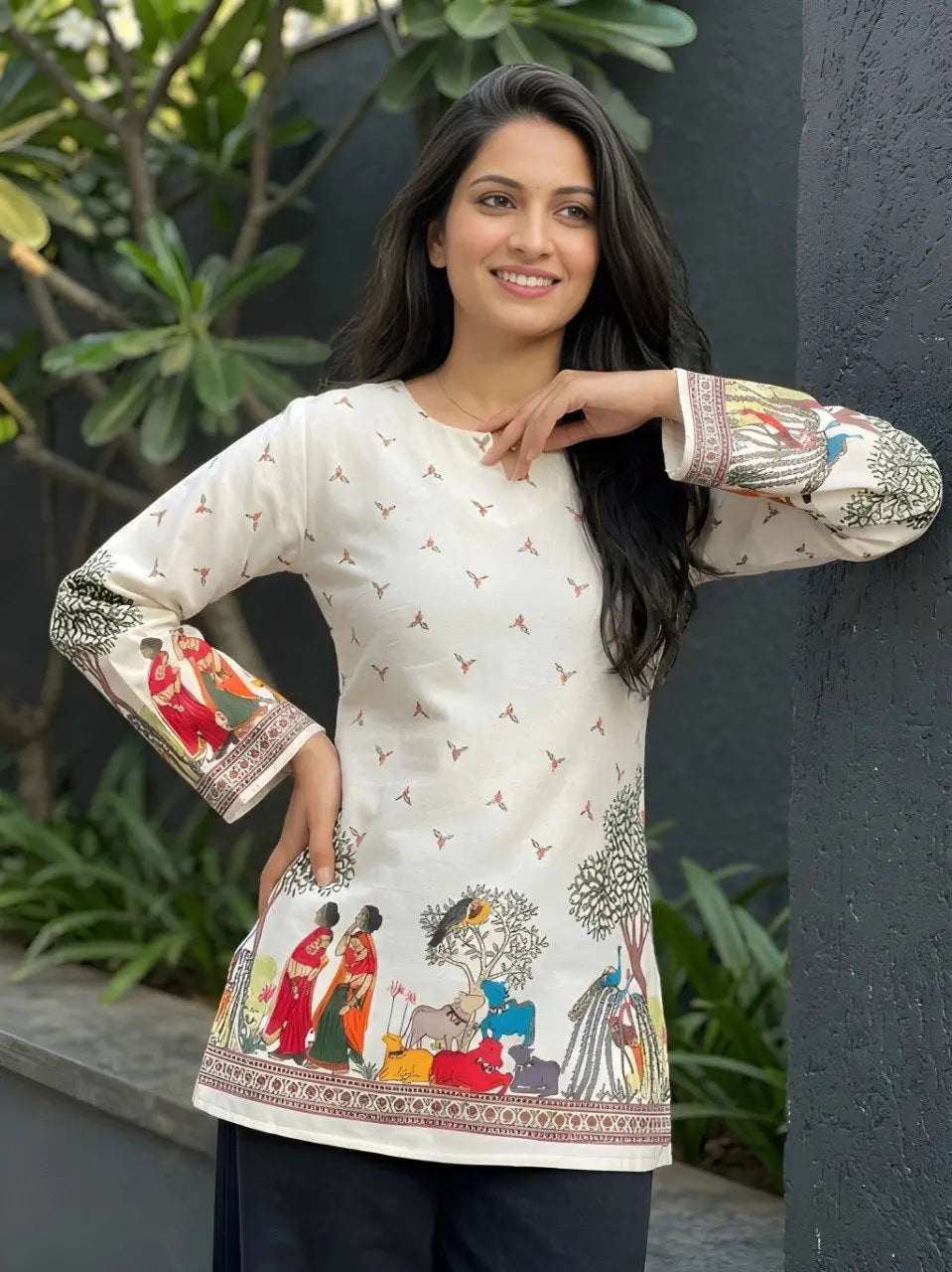 Woman wearing a white traditional outfit with colorful embroidery, standing against a dark wall with greenery.
