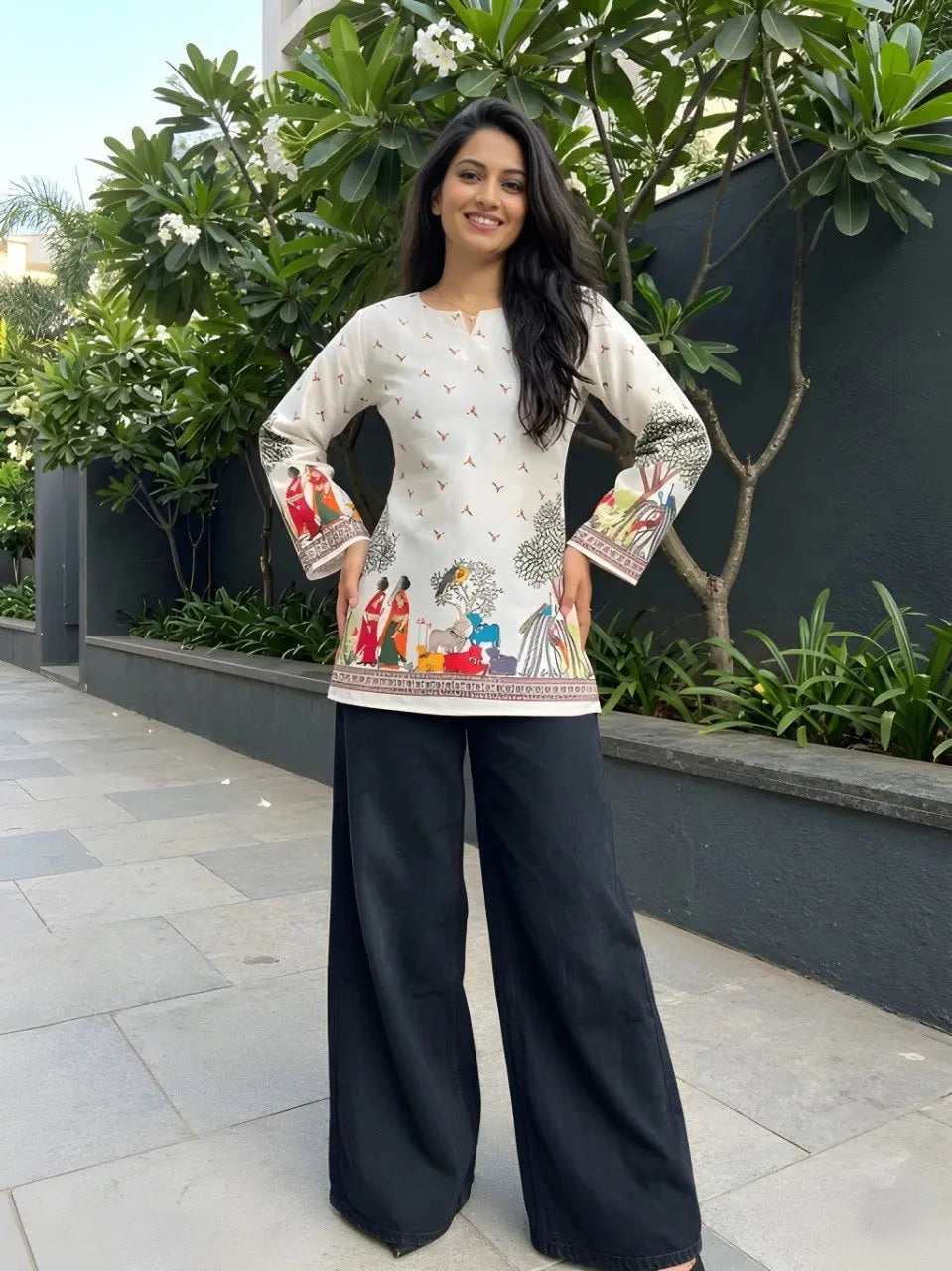 Woman wearing a traditional outfit with a patterned top and black pants, standing outdoors.