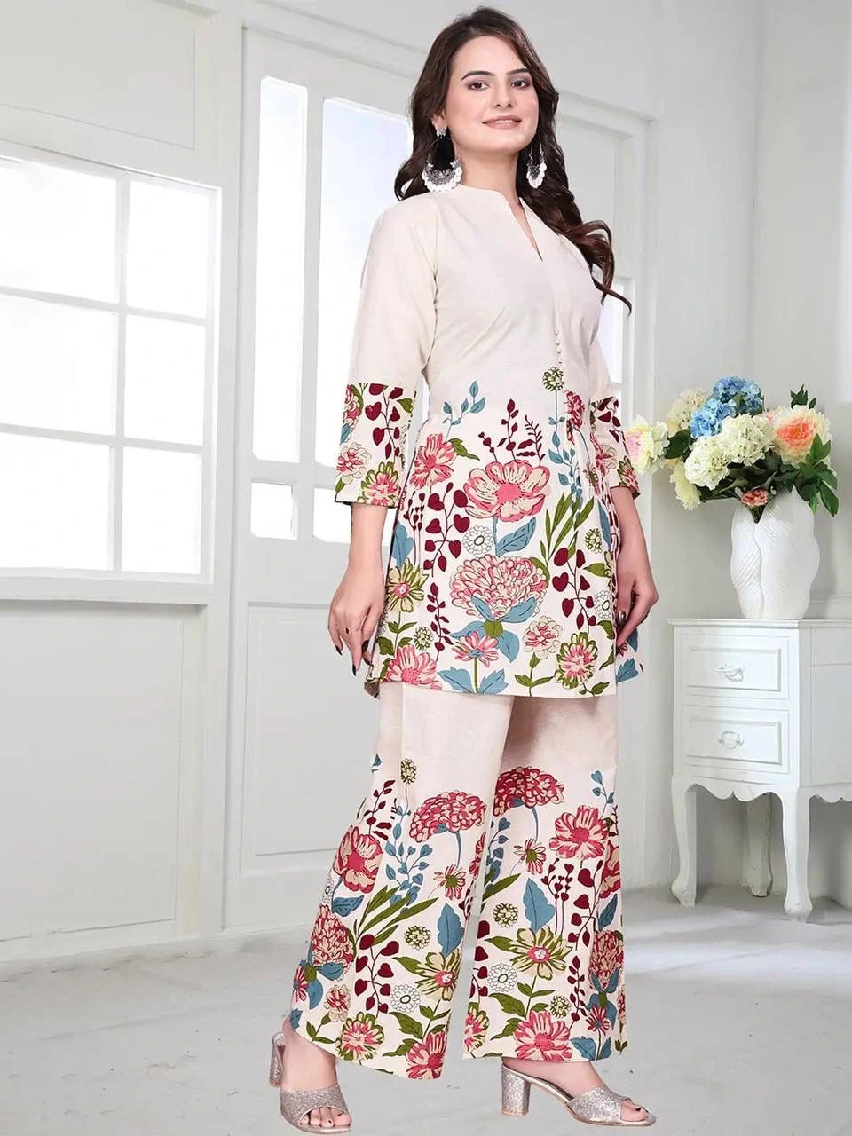 Woman wearing a floral dress in a bright room with a vase of flowers on a table.