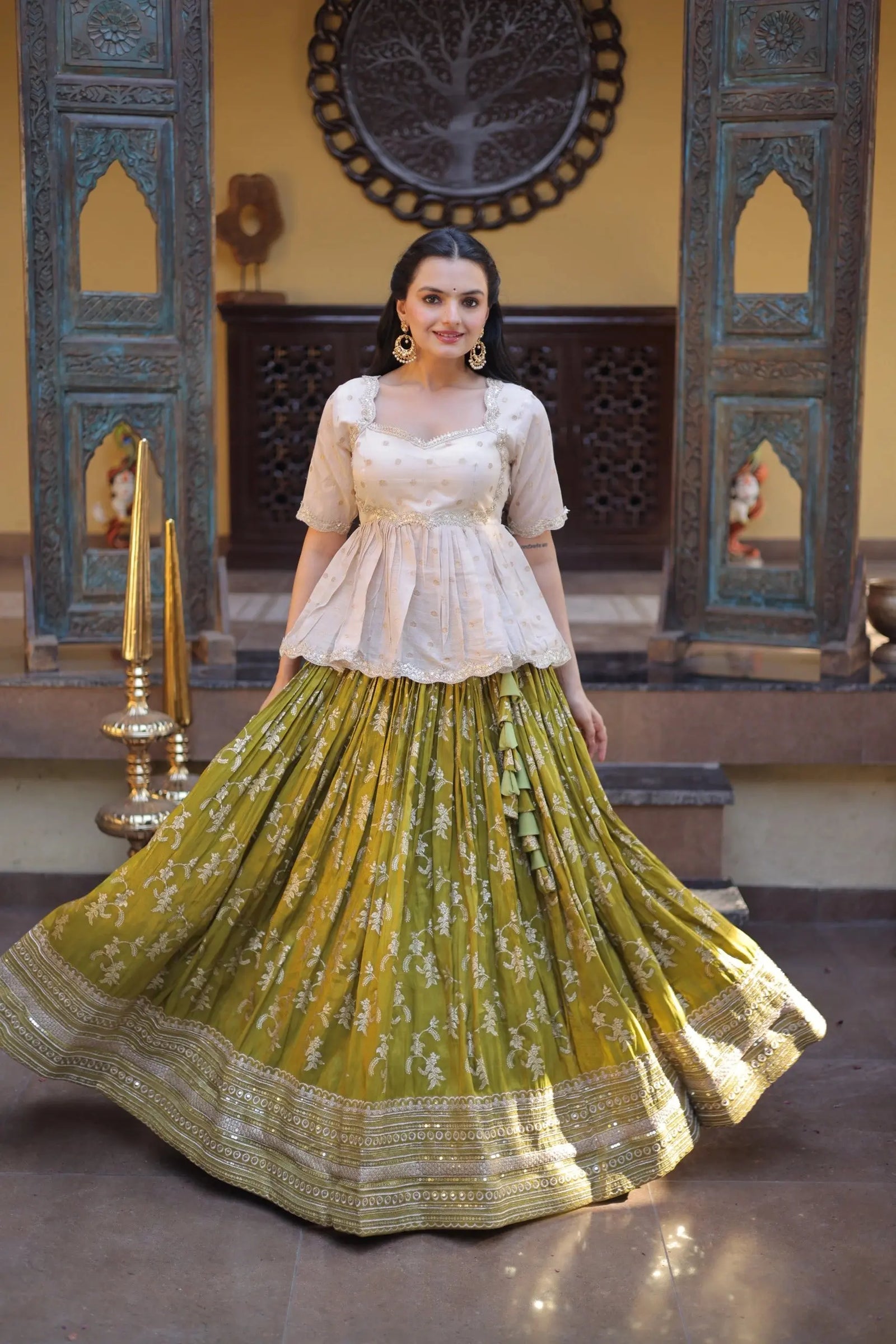 Woman in a traditional outfit with a green and gold skirt and white top, standing in an ornate room.