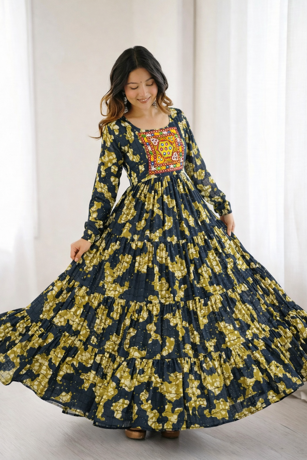 Woman wearing a long floral dress with a colorful patterned top against a white curtain background