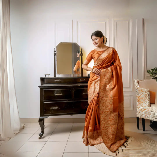 A woman models a luxurious Terracotta Orange Raw Silk Saree. The saree features a wide border and pallu with dense, tone-on-tone gold Zari weaving in a Kalamkari-style floral and paisley pattern. She wears a matching short-sleeve orange blouse and subtle gold jewelry, posing indoors next to a dark wooden vanity.