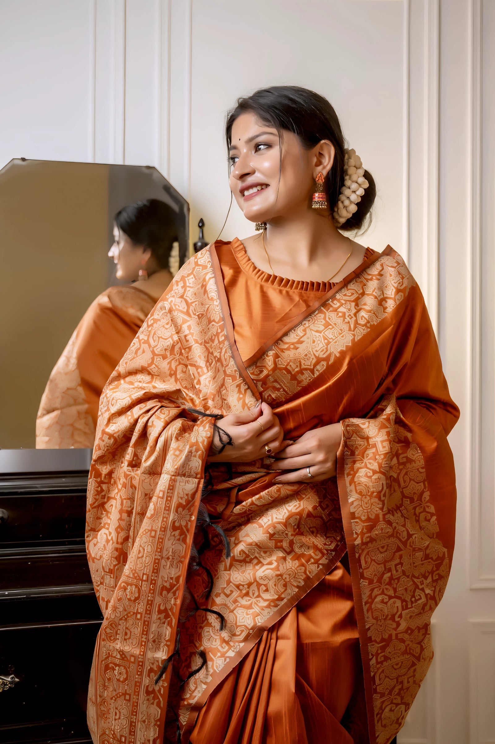 A woman models a luxurious Terracotta Orange Raw Silk Saree. The saree features a wide border and pallu with dense, tone-on-tone gold Zari weaving in a Kalamkari-style floral and paisley pattern. She wears a matching short-sleeve orange blouse and subtle gold jewelry, posing indoors next to a dark wooden vanity.