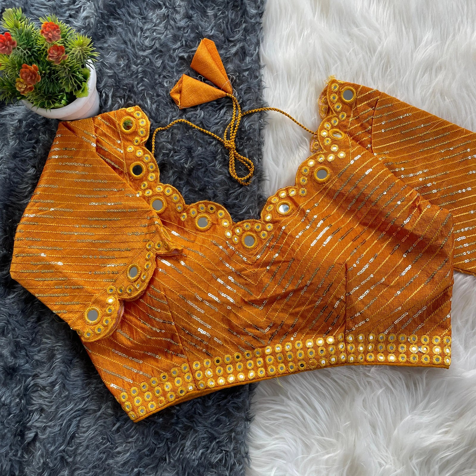 Orange bridal blouse with scallop sleeves, featuring circular mirror work and silver sequin stripes, decorative borders on sleeves and hem, and tie-up tassels, displayed on a textured grey and white fur background with a small potted plant nearby.