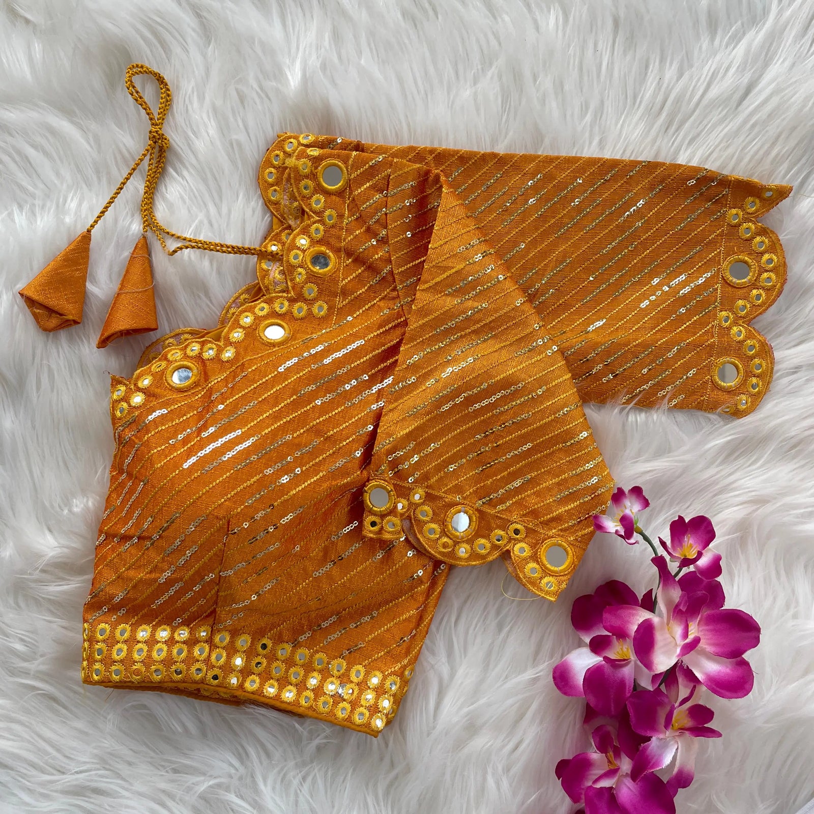 Orange bridal blouse with scallop sleeves, featuring circular mirror work and silver sequin stripes, decorative borders on sleeves and hem, and tie-up tassels, displayed on a textured grey and white fur background with a small potted plant nearby.