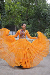 Woman in a bright orange traditional outfit with a colorful top, standing outdoors with greenery in the background.